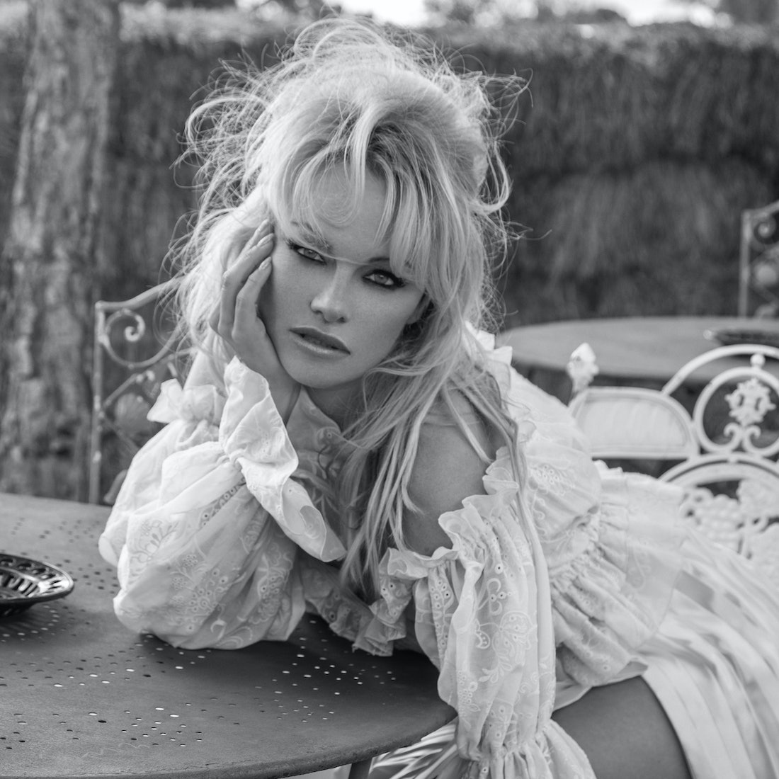 Pamela Anderson wears a white dress and sits at a table.