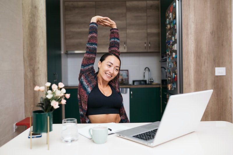 A person stretches her arms over her head while looking at her laptop at home. Stretching can help i...