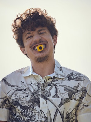 Charlie smiling with a small sunflower in his teeth wearing a white flower shirt by Erdem